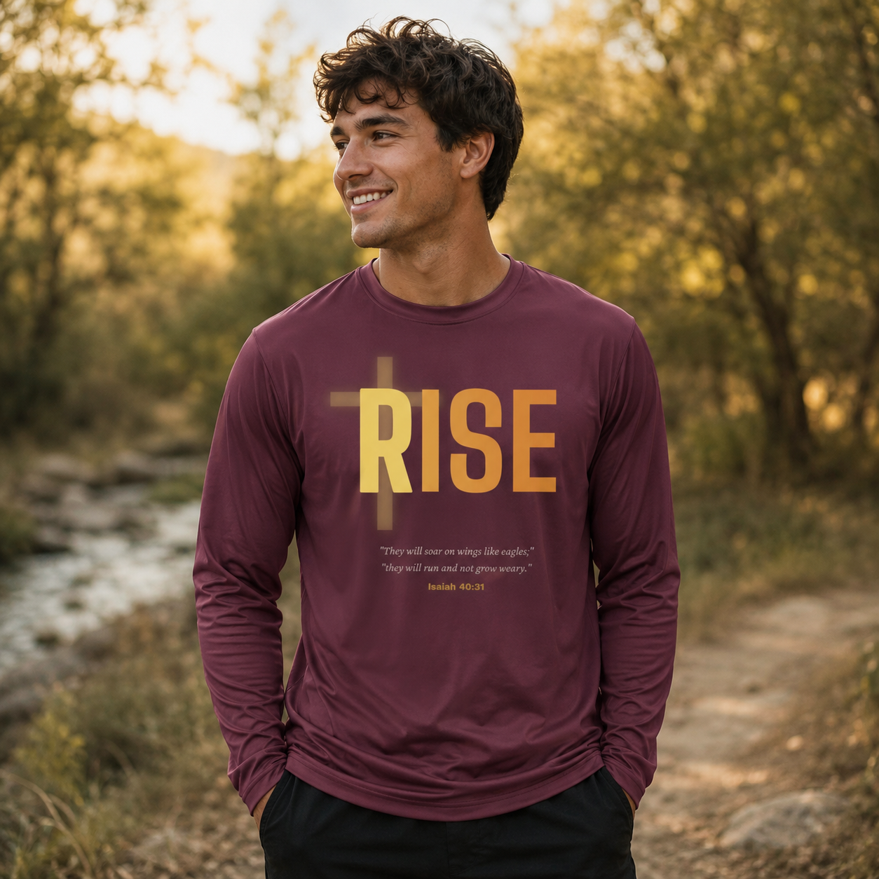 Man wearing a maroon 'RISE' shirt standing outdoors near a stream with trees in the background.
